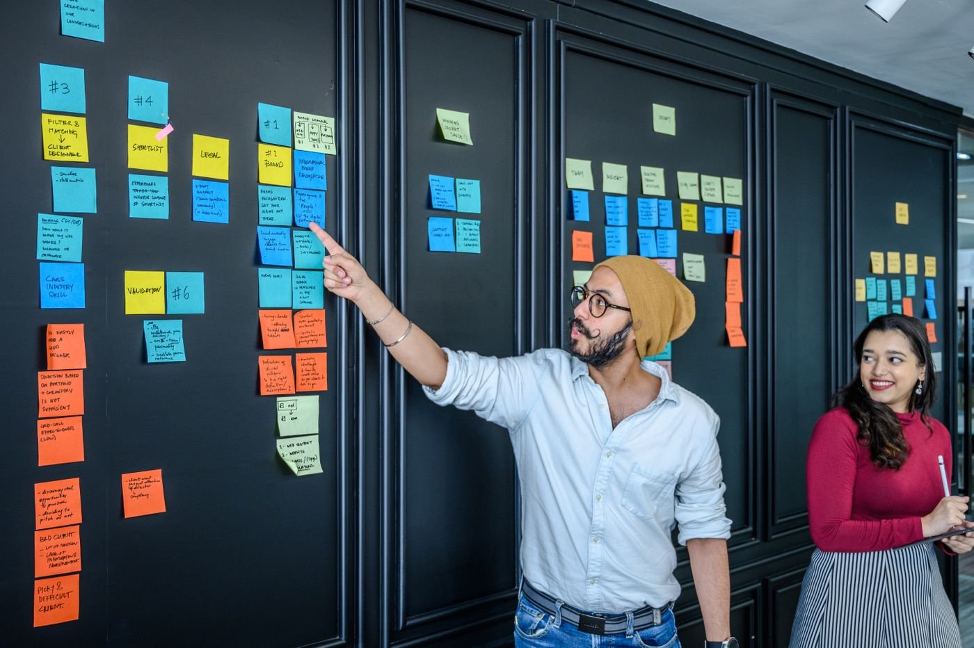 Deux collègues échangent devant un mur de post-it organisé en colonnes lors d’un point d’équipe projet
